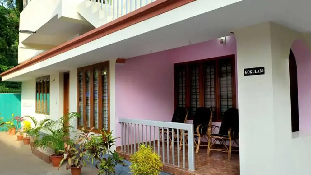 Single-story building of Gokulam Homestay Kovalam featuring a pink exterior wall, front veranda railing, and a paved courtyard.