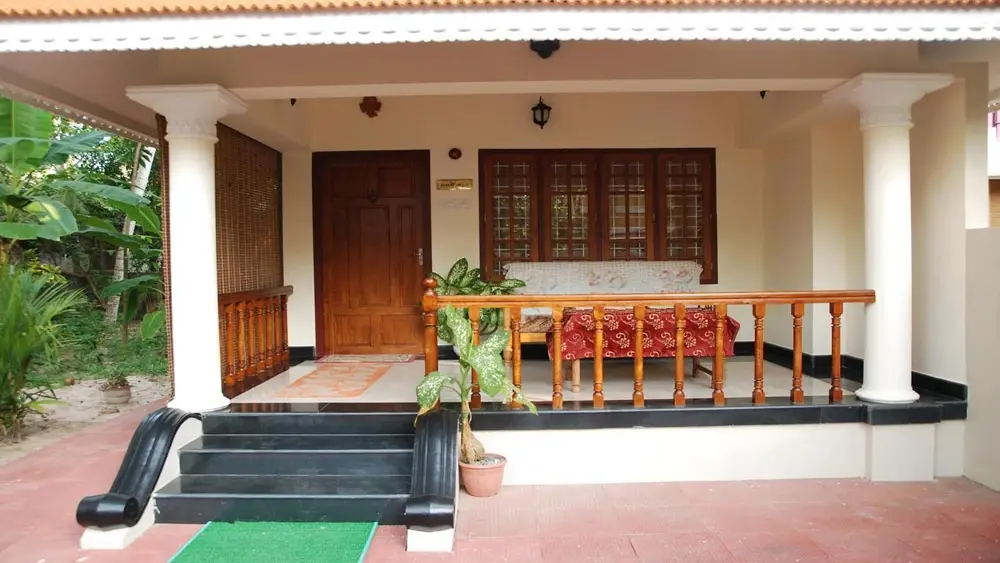 Traditional single-story building of Devikripa Homestay Kovalam featuring a wooden veranda railing, pillars, and dark stone entrance steps.