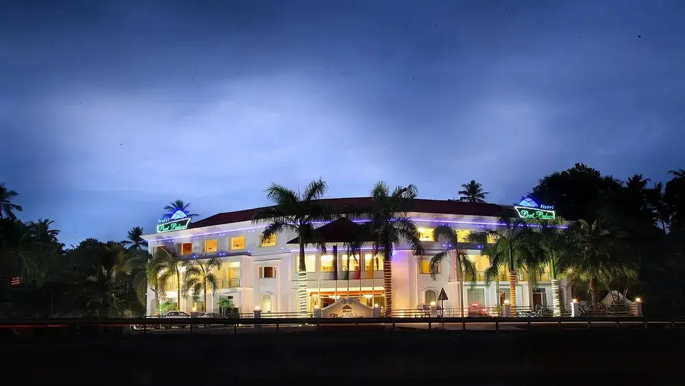 Nighttime view of the illuminated modern white building of Hotel Port Palace Kovalam surrounded by tropical palm trees.
