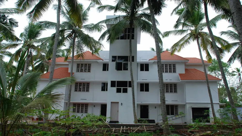 Multi-story white building of Indeevaram Apartments Kovalam with traditional tiled roofs surrounded by tall coconut trees.