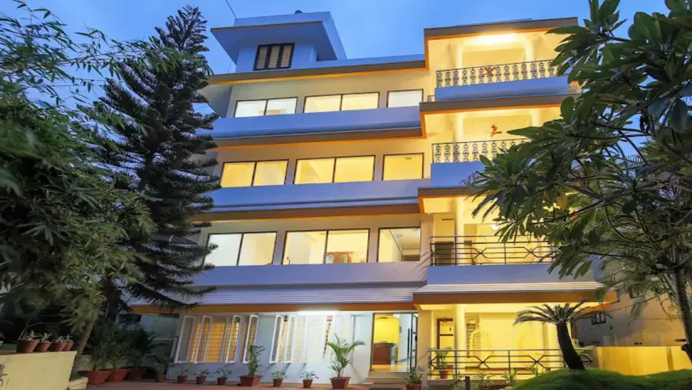 Multi-story white building of Hotel Moonlight Kovalam illuminated with warm evening lights and surrounded by tropical trees.