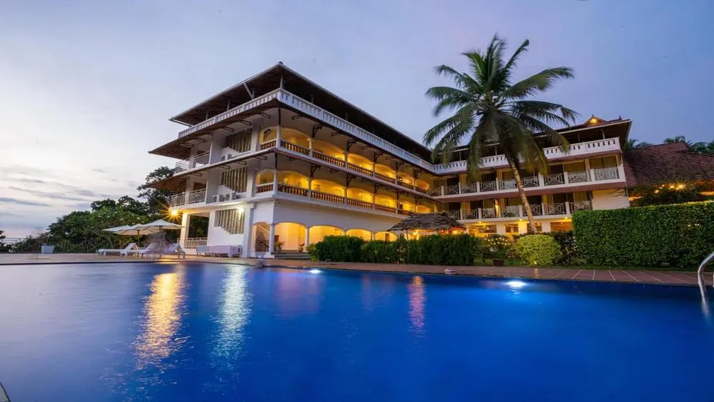 Multi-story building of Aadisaktthi Leisure Resort Kovalam illuminated with warm evening lights next to an outdoor swimming pool.