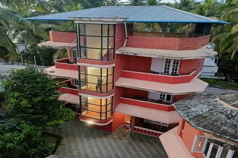 Multi-story red building of Hotel Thushara Kovalam featuring distinct circular glass windows and a tropical garden setting.