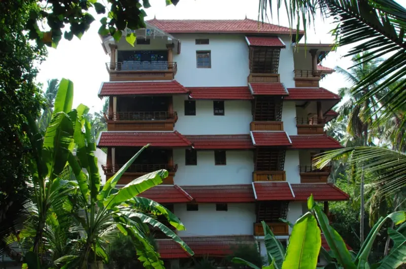 Multi-story red brick building facade of Kovalam Beach Hotel surrounded by lush green palm trees.
