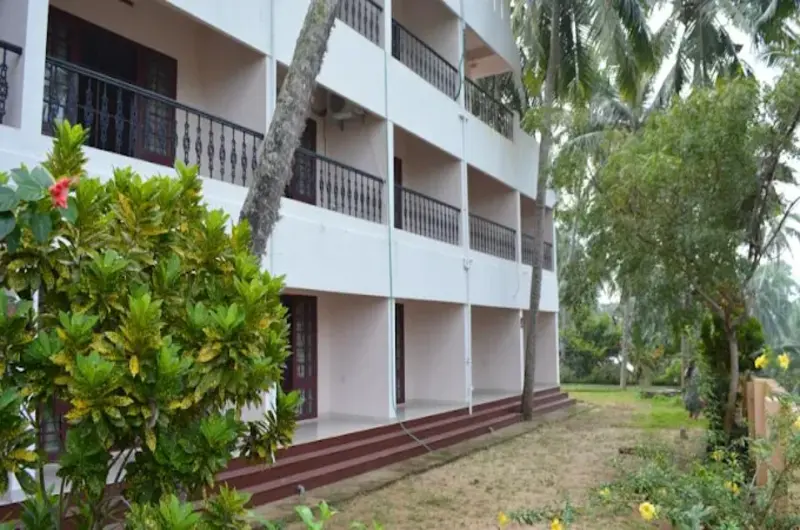 Multi-story white building of Indeevaram Apartments Kovalam surrounded by lush tropical coconut trees.