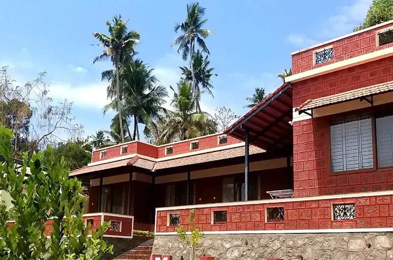 Red brick exterior facade of the homestay with distinctive large glass windows, set against a backdrop of tall coconut trees and blue skies.