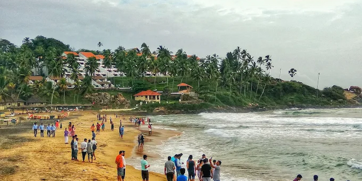 Tourists walking along Kovalam beach shoreline with waves crashing near palm groves
