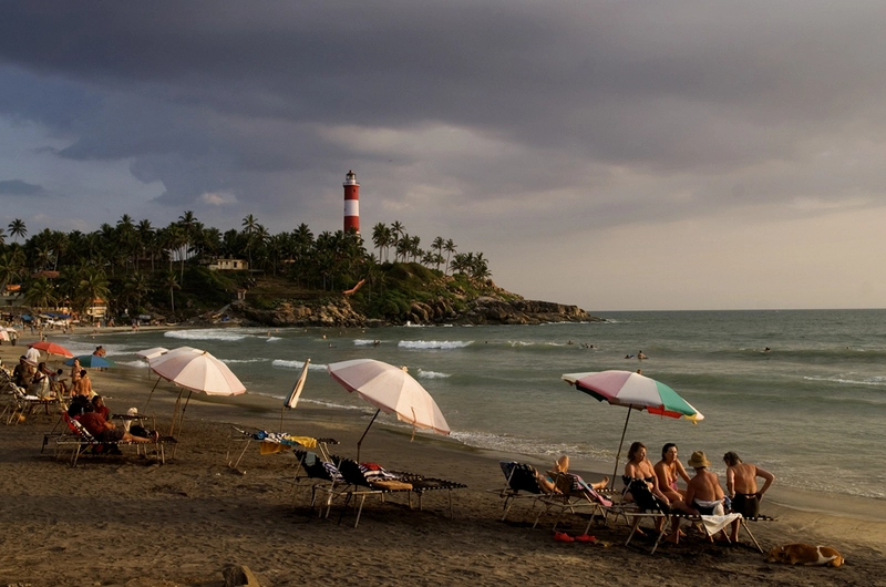 Scenic view of beachgoers sitting under colorful umbrellas on the sandy beach with the iconic red and white striped lighthouse in Kovalam Kerala